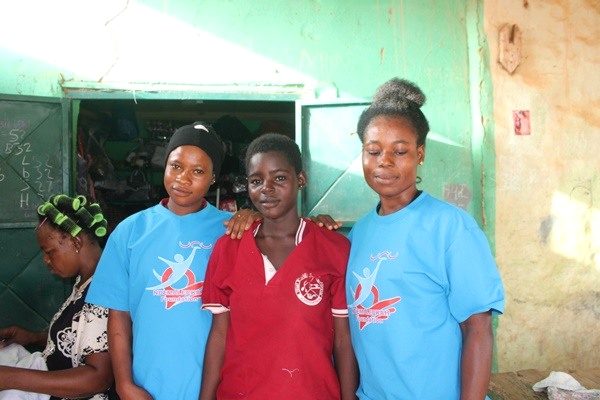 Three people stand together in a workshop, with sewing activity visible behind them.